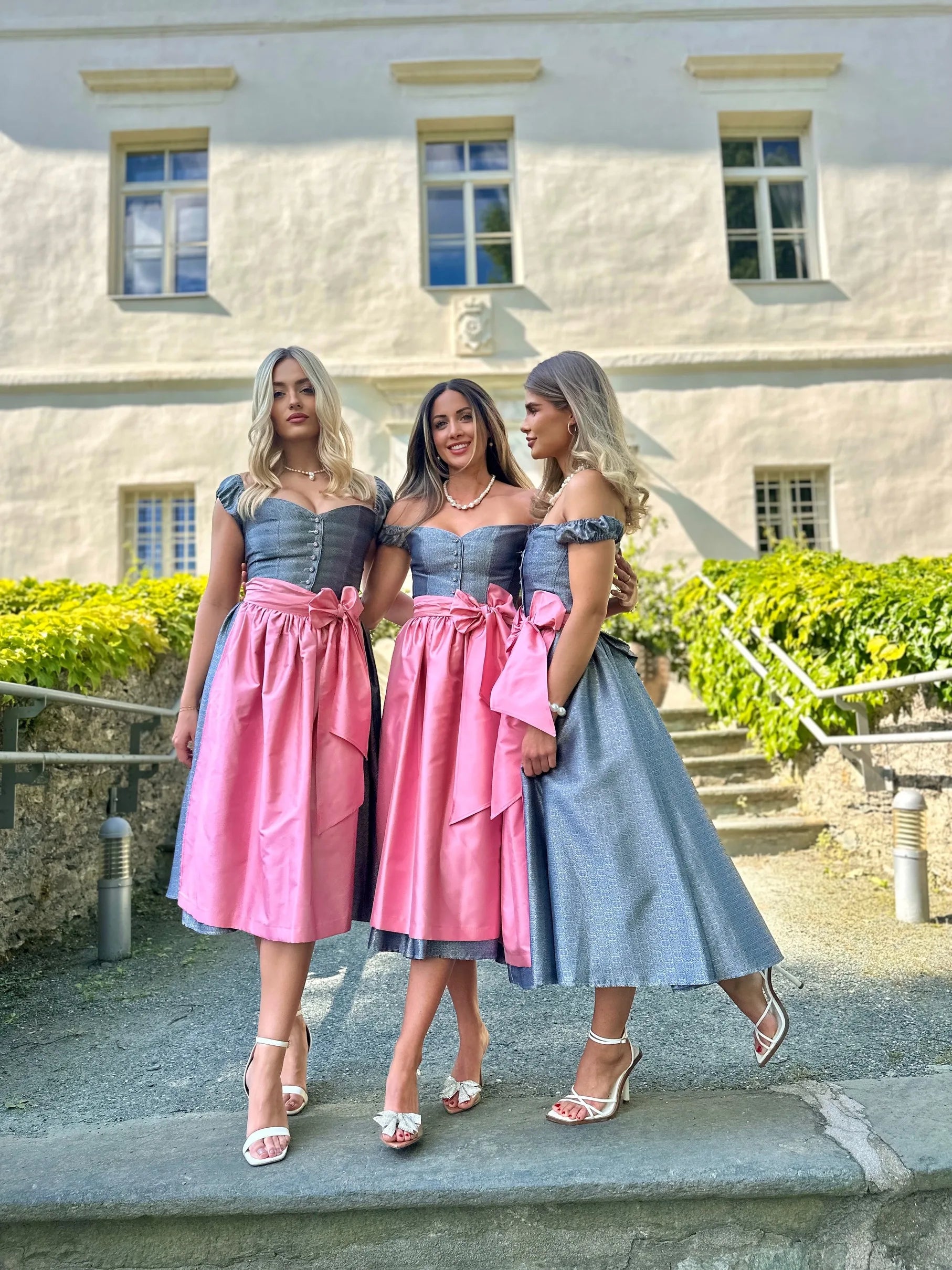 Silbergraues Dirndl Sissi Rosee Glacee mit rosa Schärpe an Frauen getragen – elegante Trachtenmode in stilvoller Präsentation.